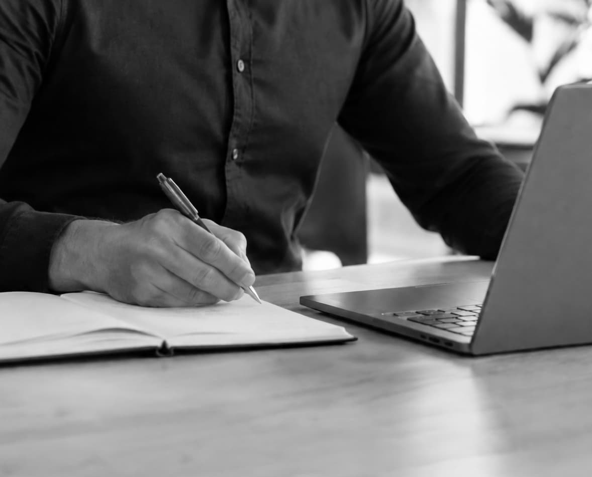 Nahaufnahme einer Person beim Schreiben von Notizen in ein Notizbuch an einem Schreibtisch mit Laptop und Unterlagen in einer hellen Büroumgebung. Das Gesicht ist nicht sichtbar.